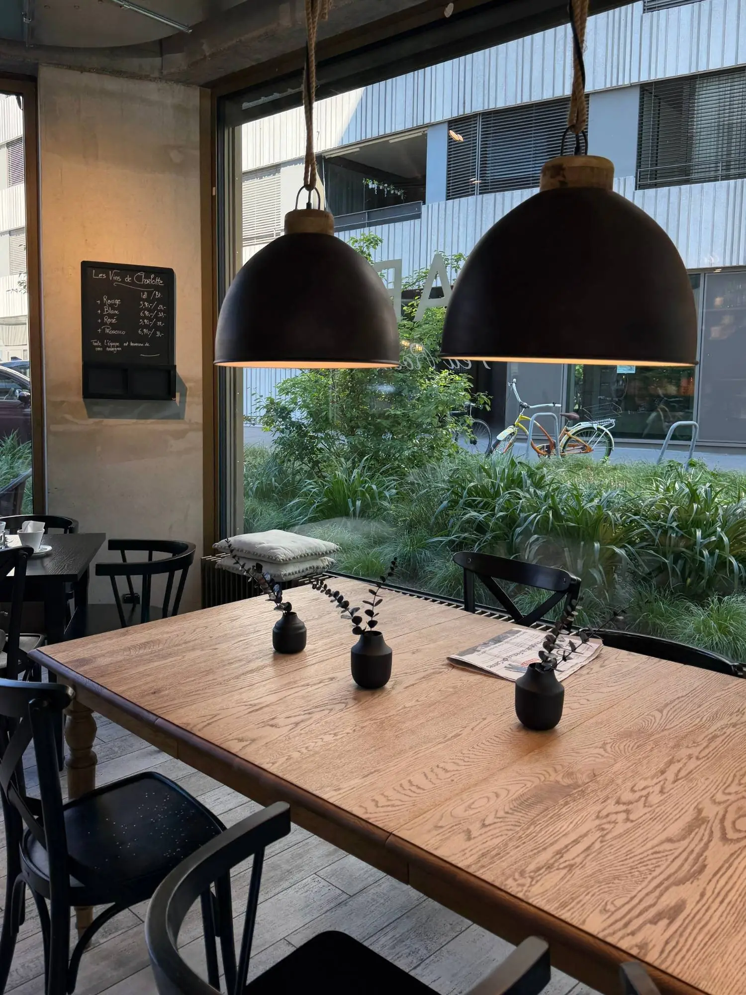 Le salon de thé : grande tablée bois clair, suspensions noires et baie vitrée sur jardin